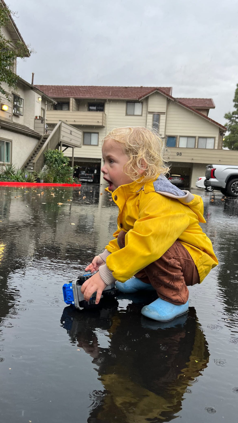 Toddler in a yellow raincoat crouched in a puddle wearing blue Meriweather sock shoes